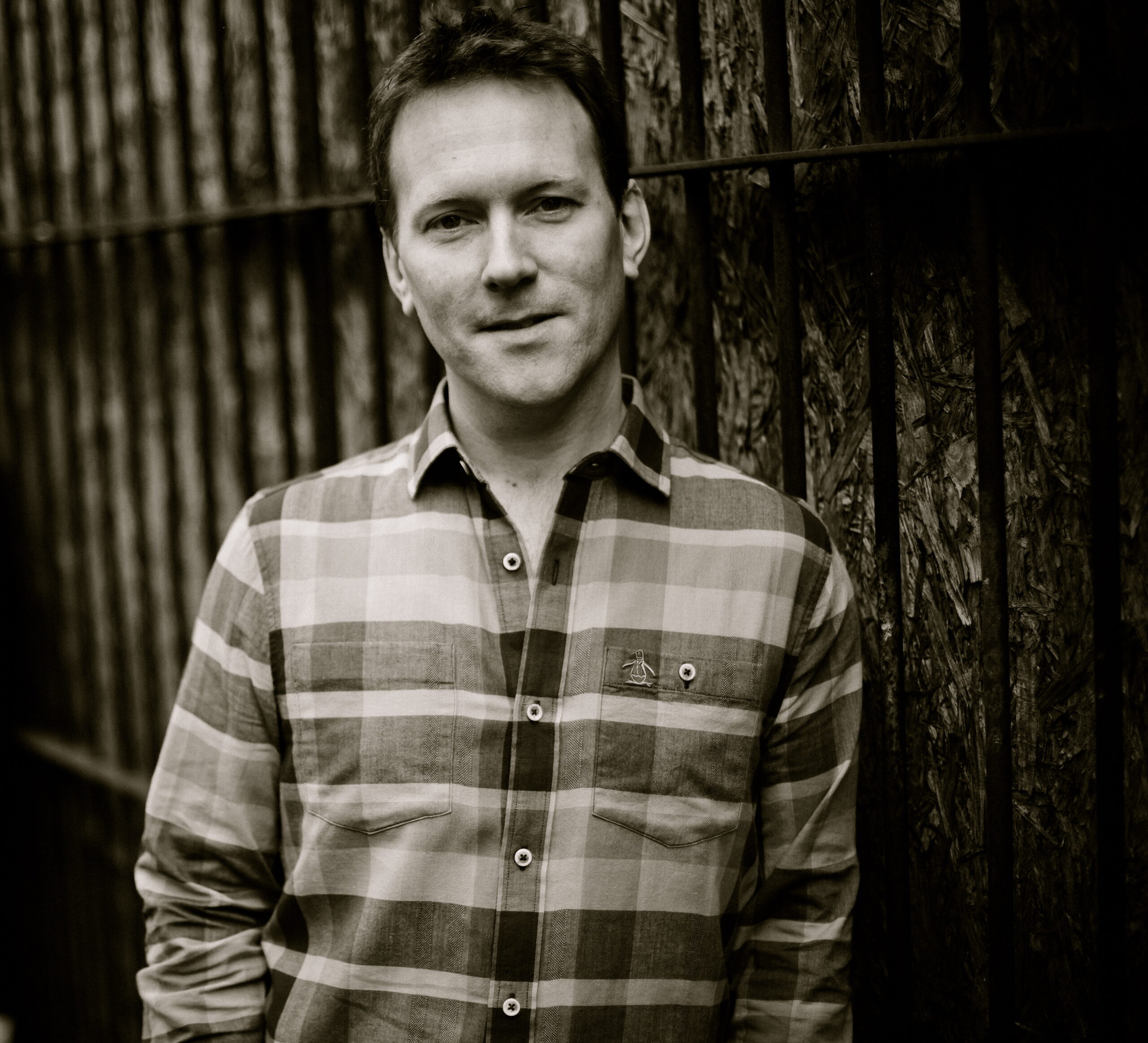 A white man in a checked shirt leans against a wooden fence.